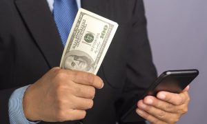 Businessman holding US cash and a phone. The PGCB fined Live! Casino Philadelphia $100,000 on Wednesday for violating the proxy wagering rule at the casino's retail sportsbook.
